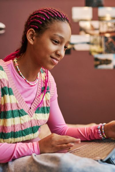 Side view portrait of black teenage girl sitting on bed and using laptop at home in pink room