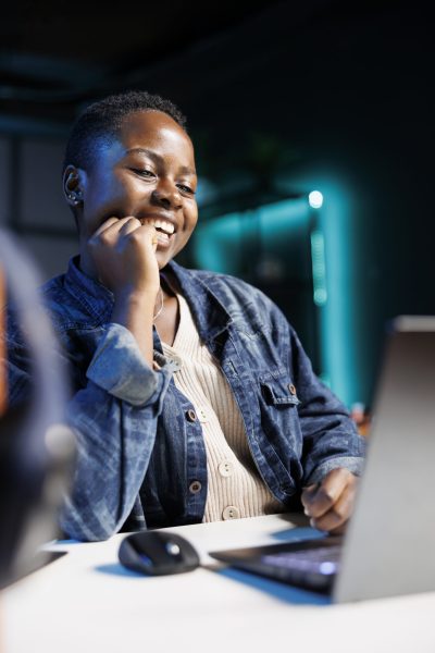 Close-up of black woman laughing while using a digital laptop at the table. African american lady with hand on her face, joyfully utilizing modern technology, browsing through her personal computer.