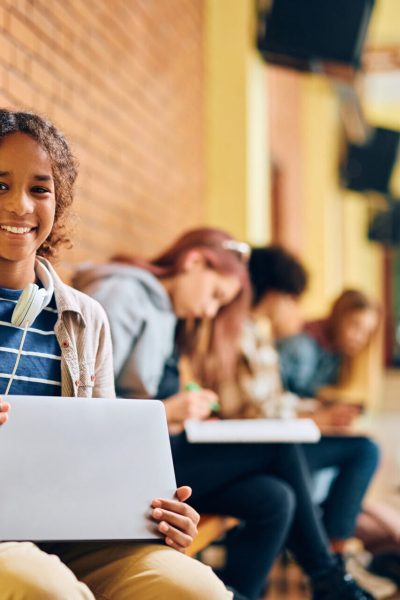Happy African American high school student learning on laptop in hallway and looking at camera. Her friends are in the background.